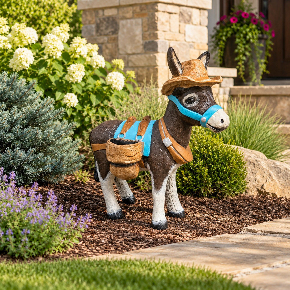 A decorative donkey garden statue wearing a brown hat, blue bridle, and tan harness with a small basket, displayed in a landscaped garden bed with mulch, shrubs, and a stone building in the background.