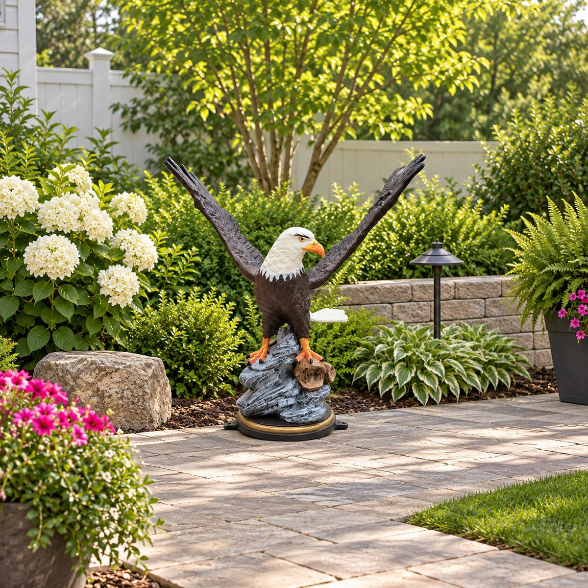 A bald eagle garden statue perched on a rock base, displayed in a landscaped backyard patio setting with greenery, flowers, and a stone wall.
