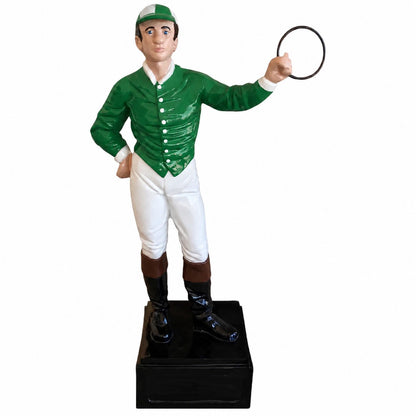 A glossy ceramic figurine of a jockey dressed in a green jacket, white breeches, and black boots, standing on a black base and holding a riding crop against a plain white background.