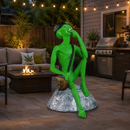 Cast aluminum green alien with beanie and smoke bag sitting on a rock and smoking.