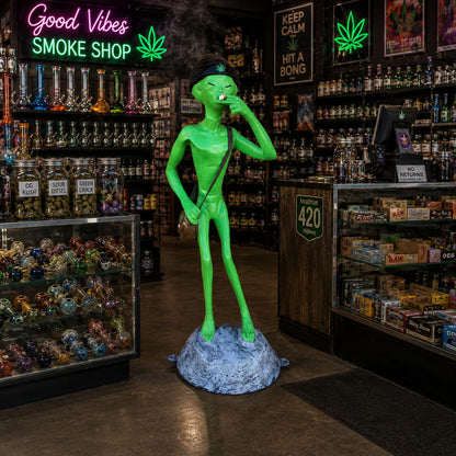 Cast aluminum green alien statue wearing beanie, holding joint, standing on rock base.