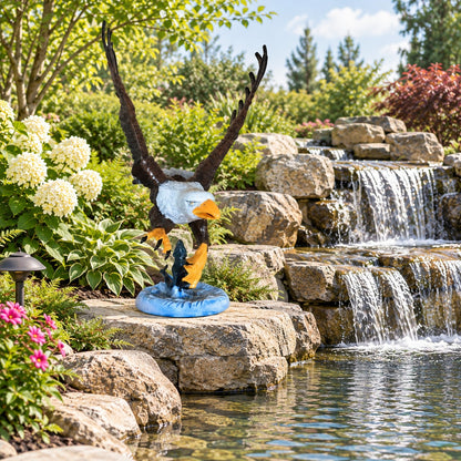 A decorative eagle statue with spread wings and a blue circular base, placed outdoors beside a stone waterfall and pond in a landscaped garden.