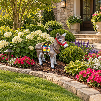 A decorative donkey garden statue wearing a colorful blanket and red saddle blanket stands in a landscaped front yard beside a stone border, flowers, shrubs, and a house entrance.