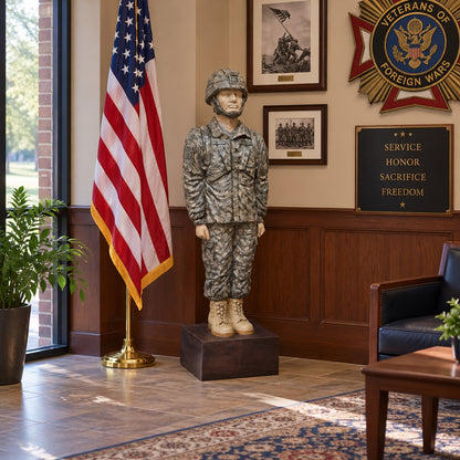 A military warrior statue in camouflage uniform stands on a dark pedestal in an indoor ceremonial room with an American flag, framed wall art, a Veterans of Foreign Wars emblem, and a service plaque.